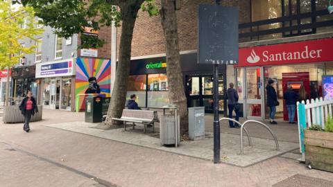 An area of a pedestrianised high street will a number of shops, seating, bins and a colourful mural