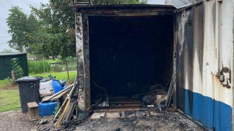 The burnt out entry to a metal container with debris inside and out. There is a plastic bin to the left.