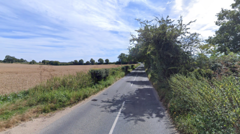 Buxton Road in Brampton, Norfolk. To the left of the single carriageway road, is a field and on the right is overgrown hedgerows and bushes. 