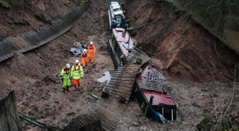 A muddy trench with three narrow boats in the mud and four people in reflective jackets