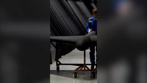 A still of the video, showing a man in blue uniform guiding a large black drone on wheels.