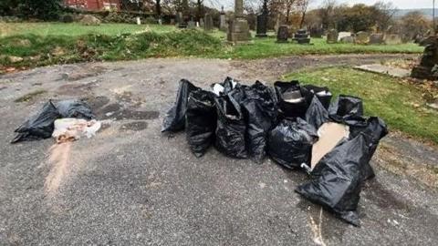 Several black bags filled with rubbish and dumped on a path in a cemetery.