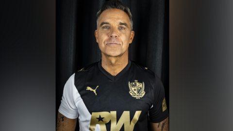 Robbie Williams, a man with grew spiked-up hair, is looking directly at the camera. He is standing against a black background, wearing a black white football shirt with the club logo and the letters RW in the centre in gold lettering.