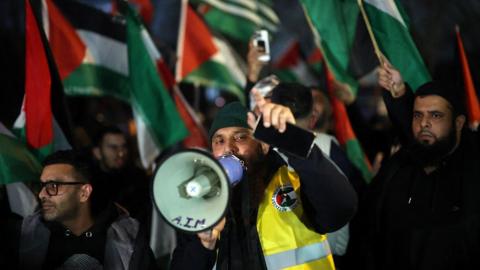Pro-Palestinian protesters gather outside the stadium before the match. A man with a hi-vis jacket has a loudspeaker. The group are carrying flags.