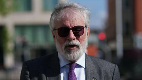 Gerry Adams, wearing a grey suit jacket, a light coloured collared shirt and purple tie. He has grey hair and a grey beard and wears dark sunglasses. He is walking on the street outside of the High Court, but the background behind him is blurred.