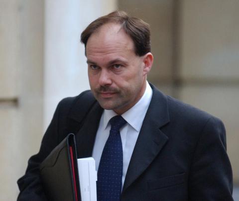 A man with thinning hair and a moustache is walking outside a court building. He is wearing a black suit and tie with a white shirt. He is holding papers contained in a black leather folder.