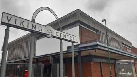 A metal sign with a white plaque reads 'VIKING CENTRE'. It stands next to an old red brick brutalist-style building. There is a BT telephone box which has been vandalised next to it. 