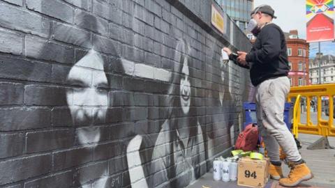 Mr Murals working on his mural in Birmingham. He is wearing a mask and side on to the camera, to the right on the photo.
