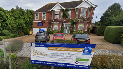 A detached brick house used as a care home, with cars parked in a forecourt. A roadside poster attached to a fence carries the words "award winning care home" and "Inspected and rated good - Care Quality Commission"