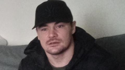 Joshua Norman wears a black baseball cap and dark jacket and top, sitting on a sofa.