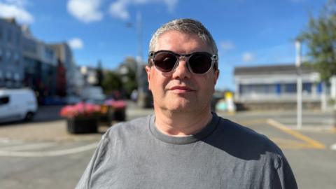 Pete Torode is a man with short grey hair look at the camera. He is wearing a grey T-shirt, and also sunglasses with tiny cameras at the corners of the lenses.