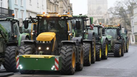 A queue of green and yellow tractors makes its way through an Oxford city centre road, with historic buildings in the background, under a misty sky