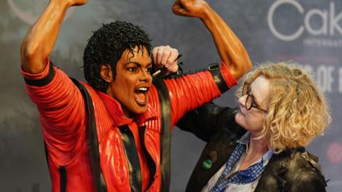 A woman with long blonde curly hair, wearing a black jacket with a white shirt and blue tie, paints the final touches on a life-size Michael Jackson cake. Jackson has been recreated in his red Thriller outfit and is holding his arms in the air with his mouth open.