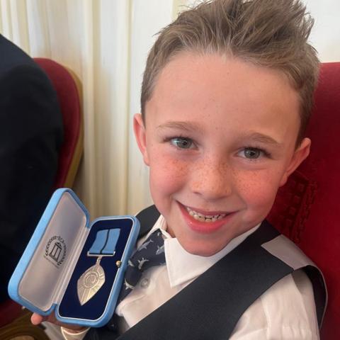 Teddy a young boy wearing a waistcoat, shirt and tie holding a medal - he is looking at the camera and smiling.