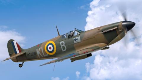 BBMF Spitfire P7350 flying across a blue sky with white clouds. It is side on and painted in camouflage colours with a yellow, blue, white and red circle on its die. There is one pilot in the cockpit. 