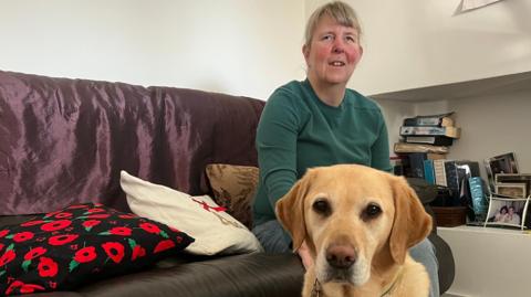Jenny sits on a sofa inside a living room, with a yellow Labrador dog sitting in the foreground. Books, framed photos and cushions are visible around them.