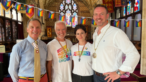 Four people smiling widely inside a church with Pride banners behind them.