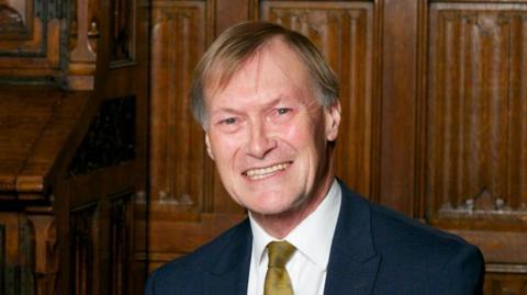 A head and shoulders image of Sir David Amess. He is wearing a navy suit jacket over a white shirt with a mustard tie. He is smiling and looking into the camera.