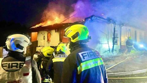 Four firefighters with helmets and firefighters uniform in front of a burning bungalow with flames emerging from the roof at night.