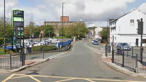 Whitworth Road with St Mary's gate retail park sign on the left and an old mill in the background along with other buildings on the right.
