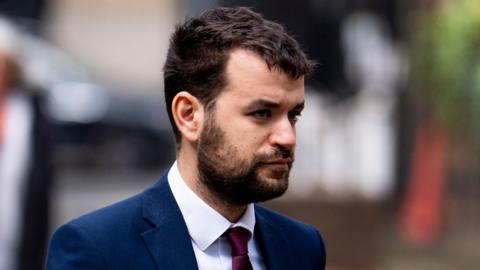 Liron Velleman arrives at Southwark Crown Court wearing a bule jacket, white shirt and purple tie