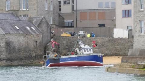A small red and blue fishing boat aground on a small beach in Lerwick. The sandy beach is surrounded by grey stone and concrete buildings. There are people in the distance overlooking the incident.