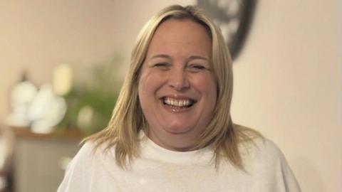 A woman is standing indoors, smiling widely at the camera and wearing a white T-shirt. The background is softly blurred, with household items and greenery, such as a clock and plants. She has blonde hair just past her shoulders.