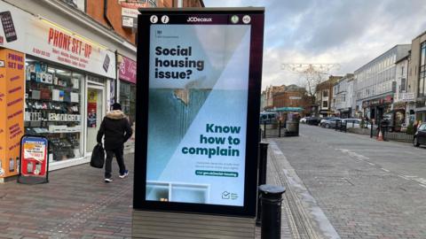 A electronic sign advertising where to complain when facing difficulties with social housing in the middle of a high street. 