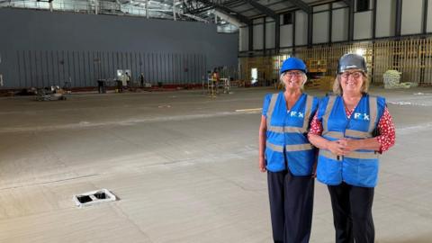 A photo of two people smiling in blue high-vis jackets. They are stood at the side of a big hallway which has works going on. They are wearing hard hats. 