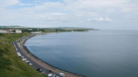 A sweeping bay, viewed from a height. A road along the bay with some caravans and other vehicles parked on it. Steep grass verges beside the road.