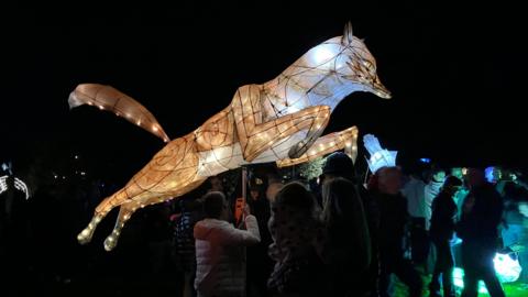 A large, glowing lantern looks like a fox mid-jump with its orange tail in the air. Someone in a white coat is holding the lantern in the air. There are crowds of people in the dark also holding other lit lanterns.
