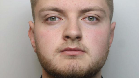 Bearded Mason Hodgson, wearing a black shirt, looks unsmiling and straight ahead in his police mugshot