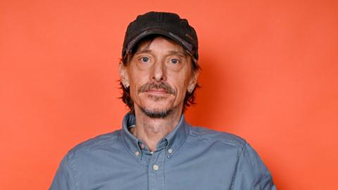 Mackenzie Crook against a red background wearing a baseball cap