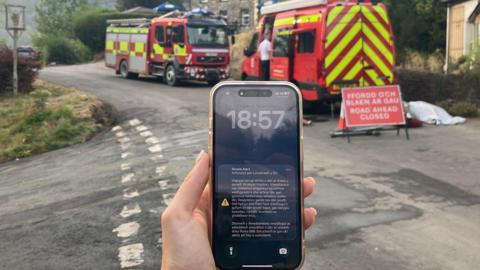 A phone screen with an emergency alert on is seen on a phone, with fire engines behind