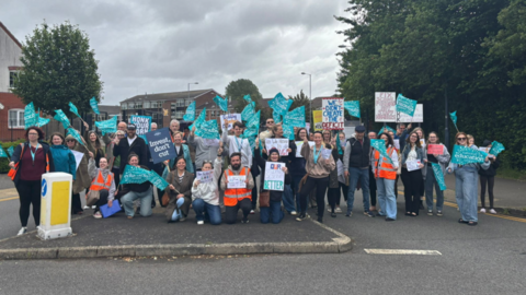 A large group of people stand across a street holding turquoise flags and protest signs with messages about education and funding. Some people kneel at the front, and a few wear high-visibility vests. The scene is set on a road with trees, houses, and a cloudy sky.