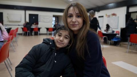 A young girl leans into a hug with her mother. Both are smiling at the camera. They are sat in a large hall surrounded by desks with orange chairs. 