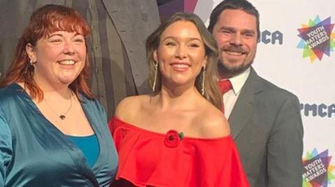 A group of people smartly dressed stand on a stage. Two women wearing dresses stand next to each other and smile. Three men dressed in suits are stood behind them. A YMCA Youth Matters Awards backdrop is behind the group.
