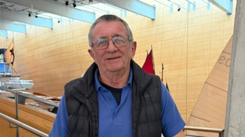 A man with short dark-grey hair, a blue polo shirt and a dark blue body warmer in a room with wooden walls