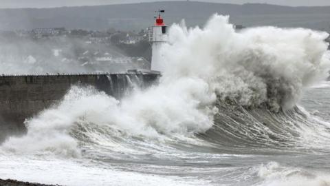 A huge foamy wave crashes into the pier at Newyln Harbour in Cornwall. The lighthouse can be seen beneath the wave, with a beach in the foreground and houses in the background