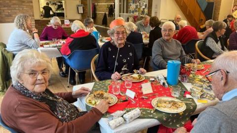 Pensioners sitting around a couple of long tables in a community centre hall. The table is set with paper Christmas table cloths, crackers and Christmas plates with turkey pie, roast potatoes, vegetables and gravy. Some people are smiling at the camera, others eating, and one woman is wearing an orange paper crown.
