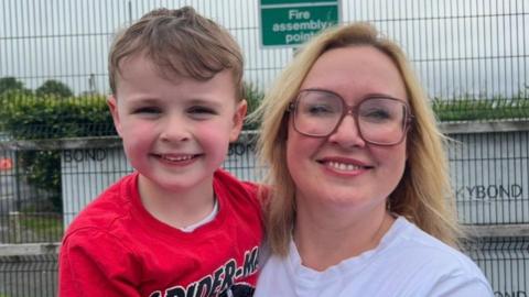 A child and a women standing looking into the camera. The boy on the left, has dark brown hair and is wearing a red t-shirt. The women on the right is wearing a white t-shirt and wearing glasses. 