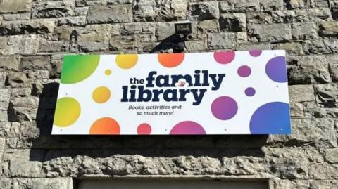 A stone wall with a sign which reads "The Family Library: books, activities and so much more!" It is white with multi-coloured spots.