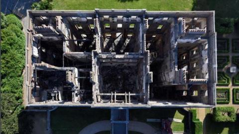 An aerial view of the house which has been burned out by fire. The outside and inside walls remain but the inner floors are gone and so is the roof. The gardens surrounding the house are green and manicured.