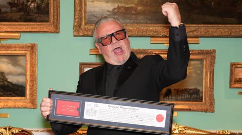 Ray Winstone pictured in a black suit and tinted glasses raises a triumphant fist while holding a framed certificate, standing in front of gold-framed paintings.