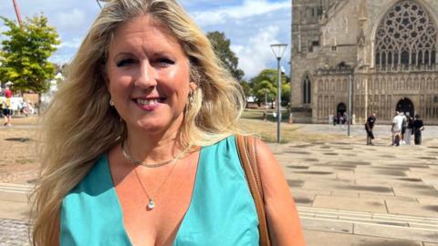 Councillor Alison Sheridan wearing a topaz summer dress with a brown leather handbag on her shoulder. She is standing in Cathedral Green in Exeter in front of the cathedral.