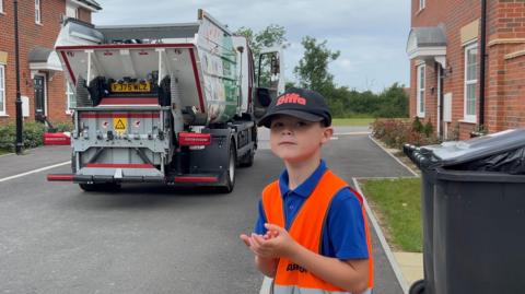 Image shows Ashton stood with orange high vis on and blue top. He has a black cap on that says 'Biffa' in red. Behind him is the food waste truck.