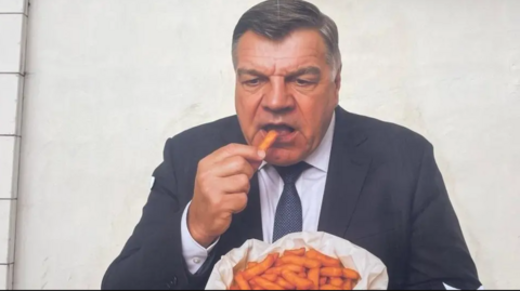 Sam is pictured in a navy suit and tie holding a bag of orange chips and putting one in his mouth with quite an intense look in his facial expression.