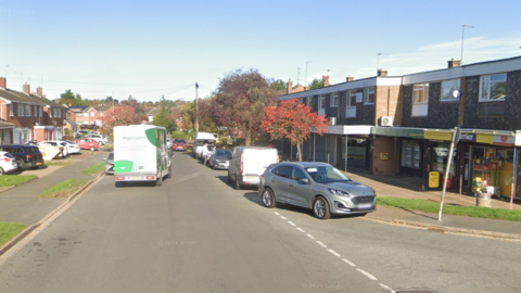 A residential street with cars parked on both sides of the road and a Morrisons van driving between them. Two convenience stores are situated next to each other. It is a sunny day with blue sky.