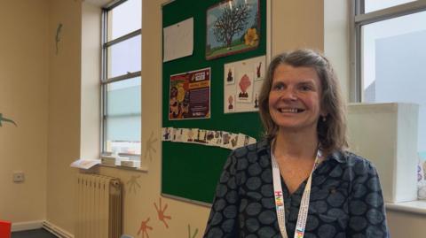 Jo Lamb is in a room which has a green noticeboard with pictures of a tree and people. The walls of the room have brightly coloured transfers in the shape of paint splodges and there are two windows.