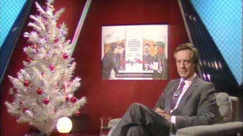 Barry Norman Sits beside a white Christmas tree and a red background with a screen, showing images from the film 'Trading Places'